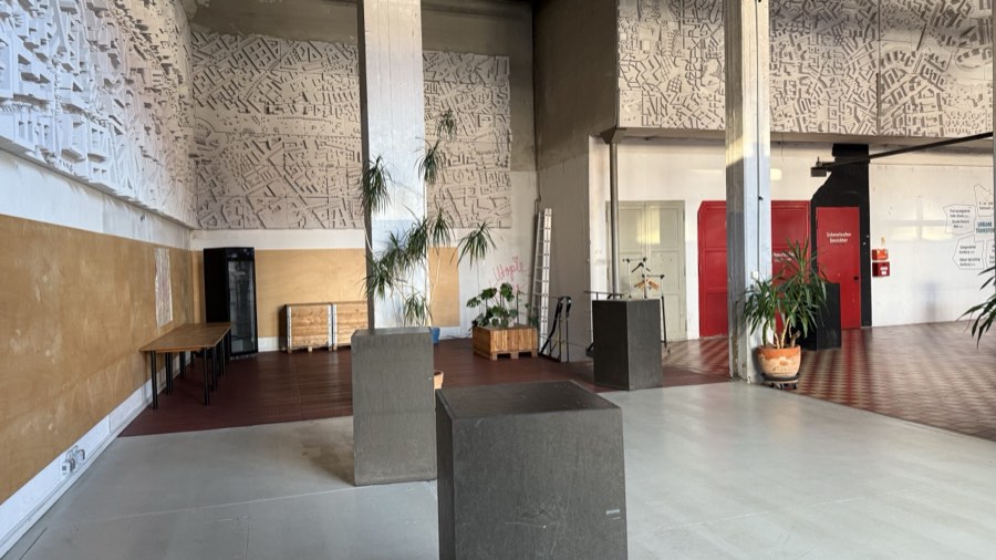 Eingangsbereich der Halle mit Betonsäulen, Pflanzen und Stadtrelief an der Wand